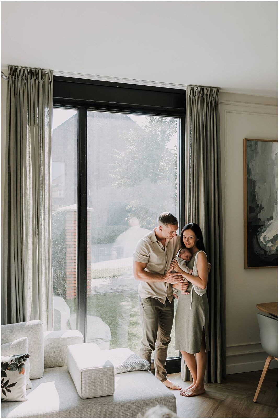 Papa en mama poseren bij het raam met hun baby tijdens een newborn fotoshoot thuis in amersfoort